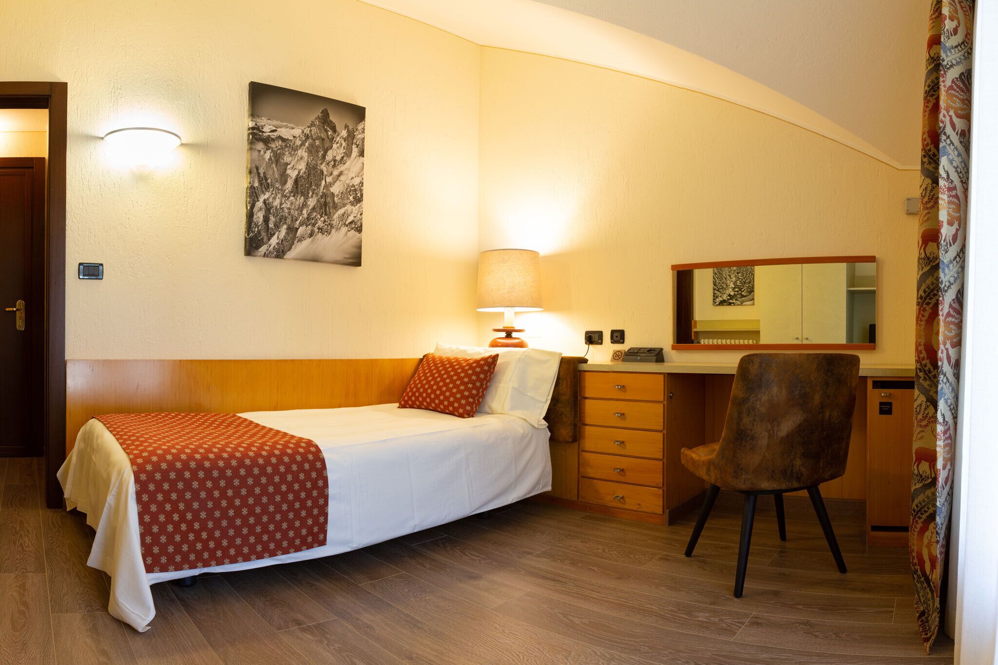 A hotel room with a bed, a desk, a chair, a lamp, a mirror, and a picture on the wall.