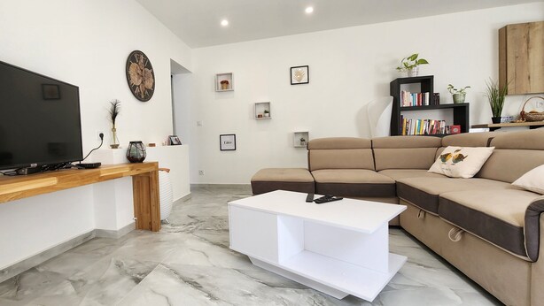 Un salon moderne avec un téléviseur à écran plat, une table basse en bois, un canapé d’angle beige et une table d’appoint blanche.