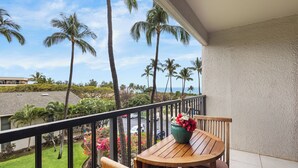 A balcony with a wooden table and chairs, overlooking a street with palm trees and a view of the ocean. - Oceanview Condo | Steps to Kamaole Beach II | Top-Floor Kihei Comfort w/ AC (Kihei)