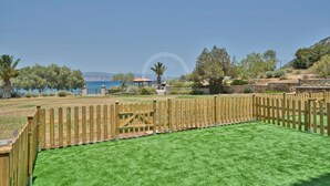 A fenced outdoor area with artificial grass, a wooden gate, and a view of a body of water and hills in the background. - Armonia Apartment in Kouverta (Ermioni)