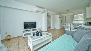 A modern living room with a flat-screen TV, a sofa, a coffee table, and a kitchen area with white cabinets and a dining set. - Armonia Apartment in Kouverta (Ermioni)
