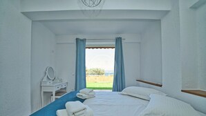 A bedroom with a bed, a bedside table with a mirror, and a view of a green landscape through a window with blue curtains. - Armonia Apartment in Kouverta (Ermioni)