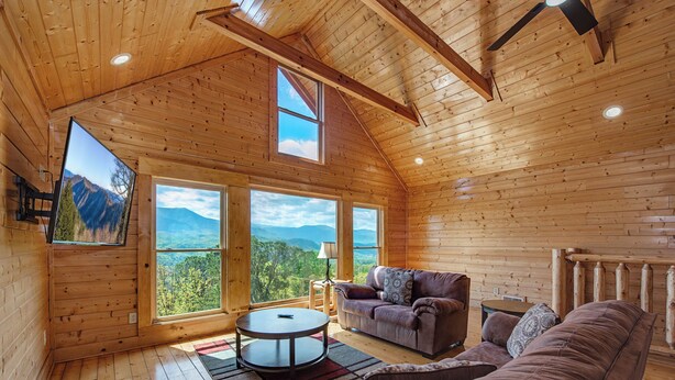 A wooden cabin interior with a sofa, a round table, and large windows offering mountain views. - Papa Bear's Cabin – 8BR with stunning Mt. LeConte views (Gatlinburg)