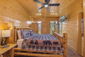 Ein Schlafzimmer einer Holzhütte mit einem Bett, einem Deckenventilator, einem Fernseher und einem Fenster.