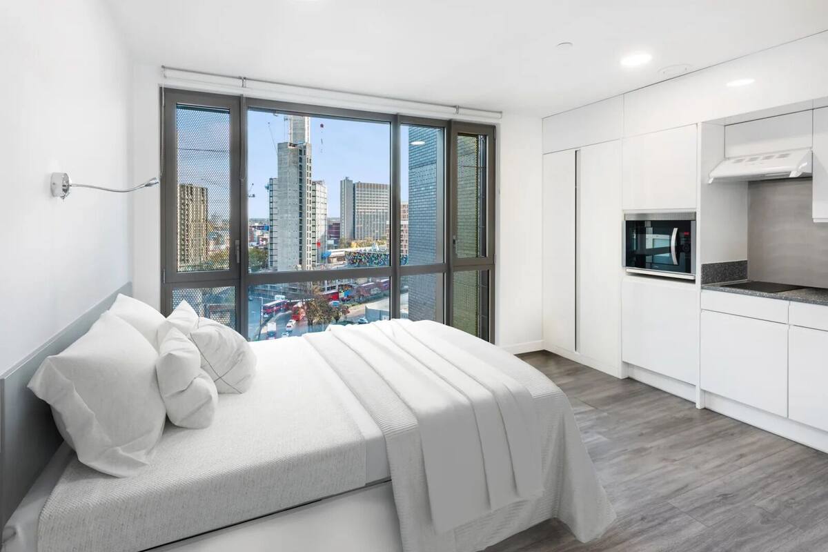 A modern bedroom with a large window, a bed, and a kitchenette.