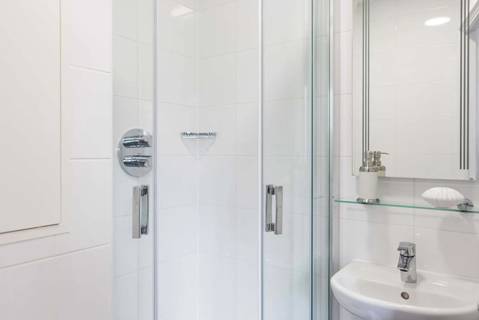 Et moderne badeværelse med en bruseniche med glasvægge, en hvid vask og en glashylde med toiletartikler.