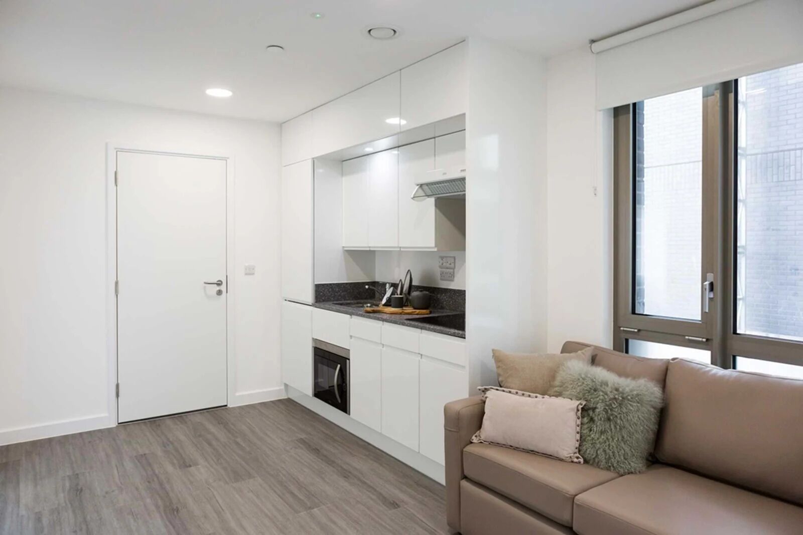 A compact kitchen with white cabinets, a black countertop, and a built-in oven. There's a beige sofa with pillows and a window with blinds.