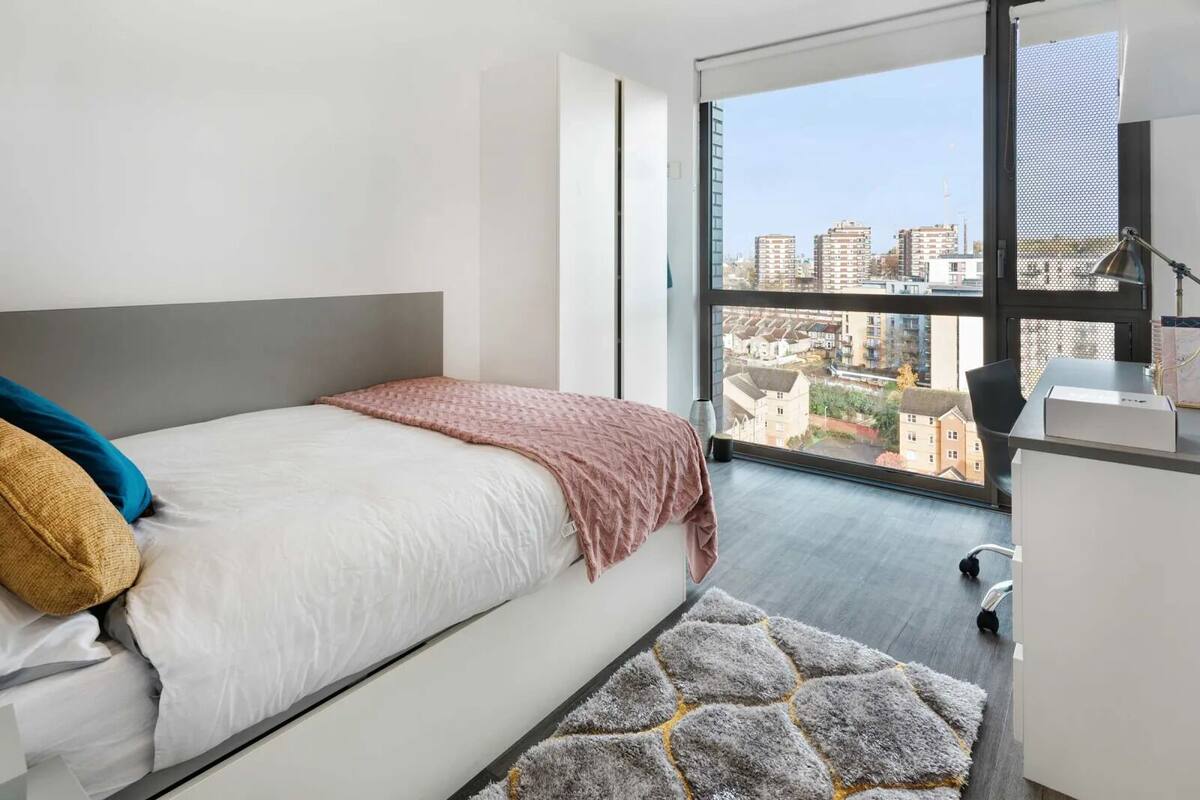 A modern bedroom with a large bed, a desk, and a view of a cityscape through a large window.