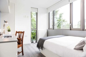 A modern bedroom with a bed, a desk, and a chair. There is a window providing natural light and a view of greenery outside. - Chapter Lewisham in London - Campus Accommodation (London)