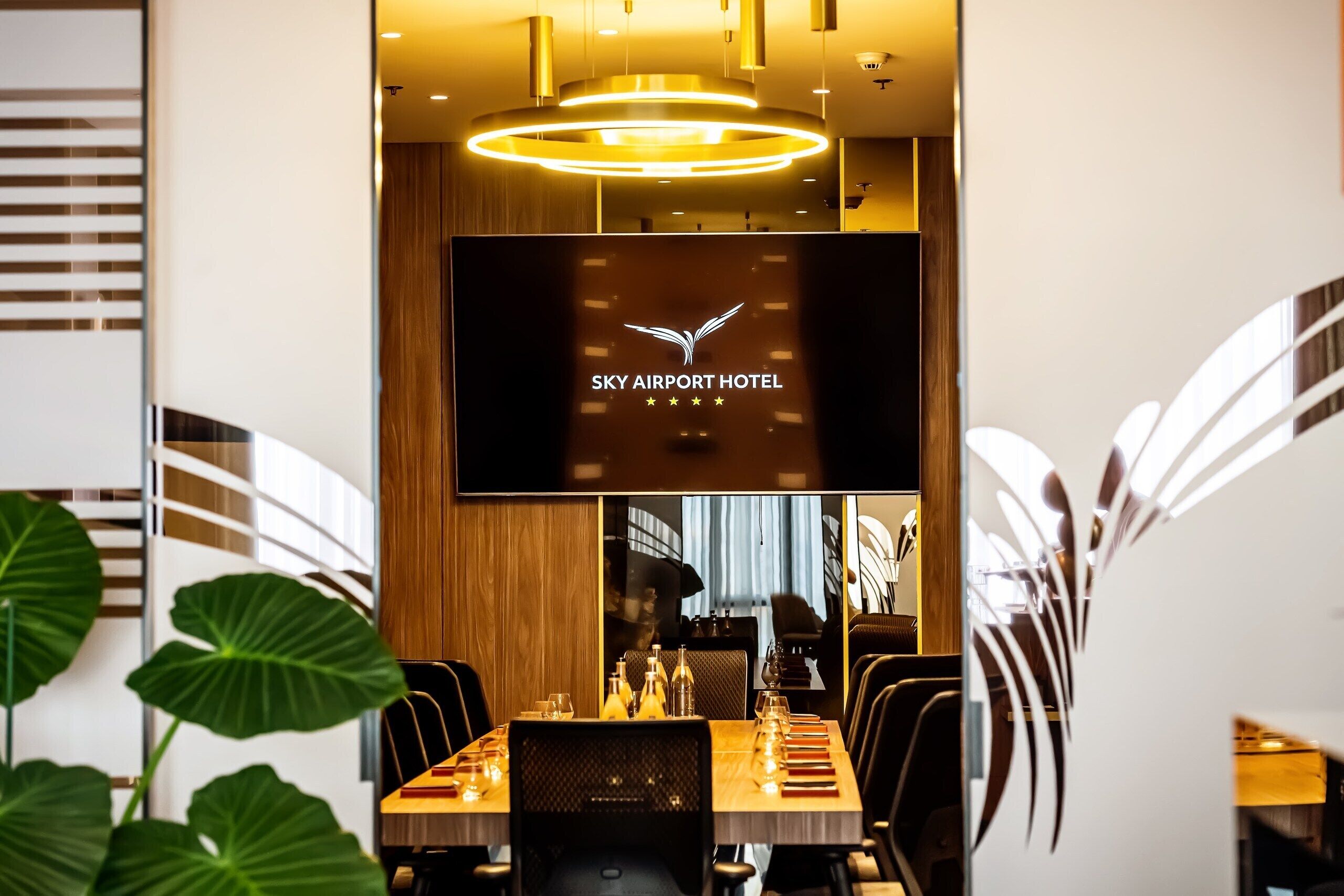 Un hall d’hôtel moderne avec un grand écran de télévision affichant le nom de l’hôtel, un espace repas avec une longue table dressée pour un repas, et une grande plante au premier plan.