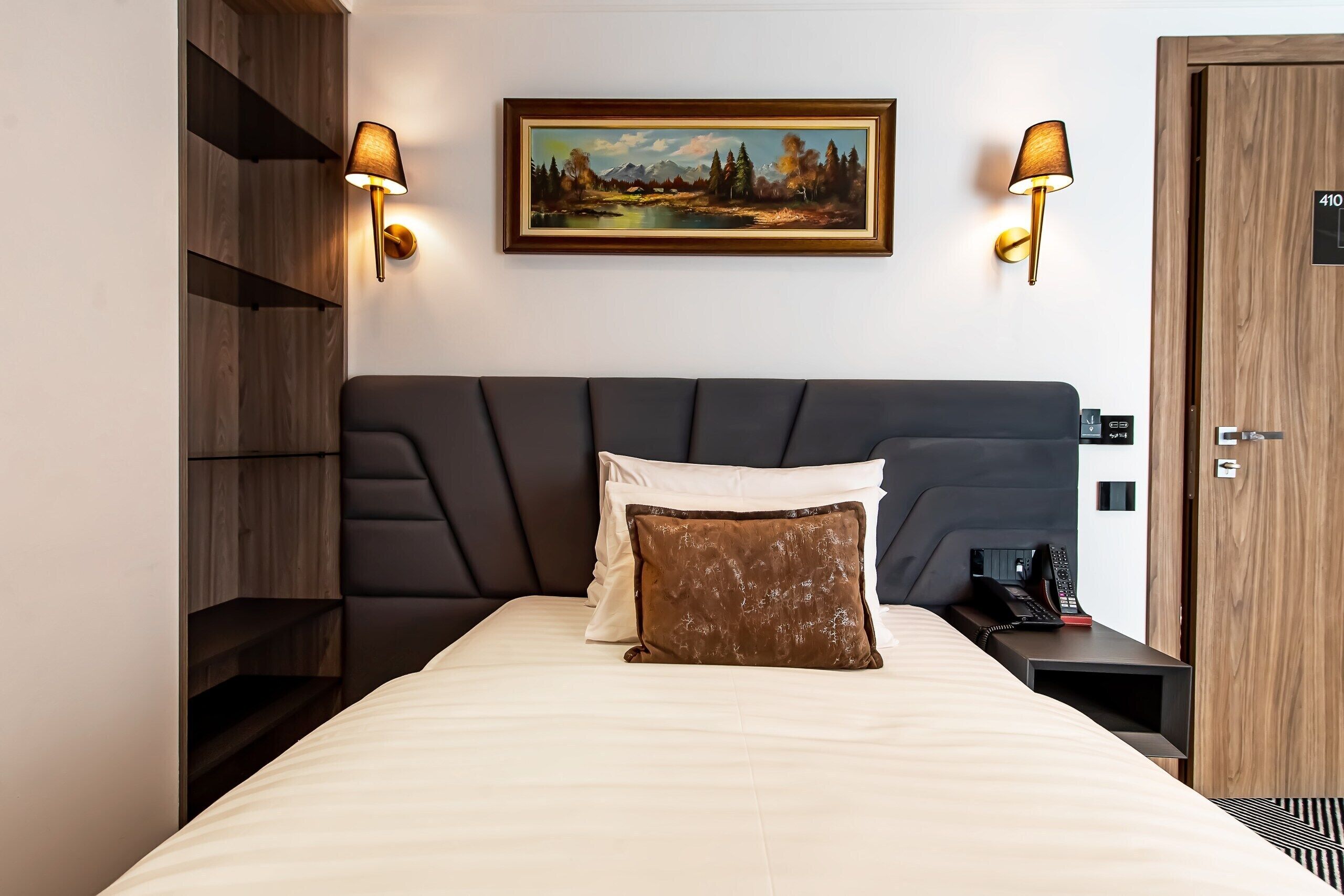 A modern hotel room with a large bed, two wall-mounted lamps, a painting, a wooden wardrobe, and a bedside telephone.