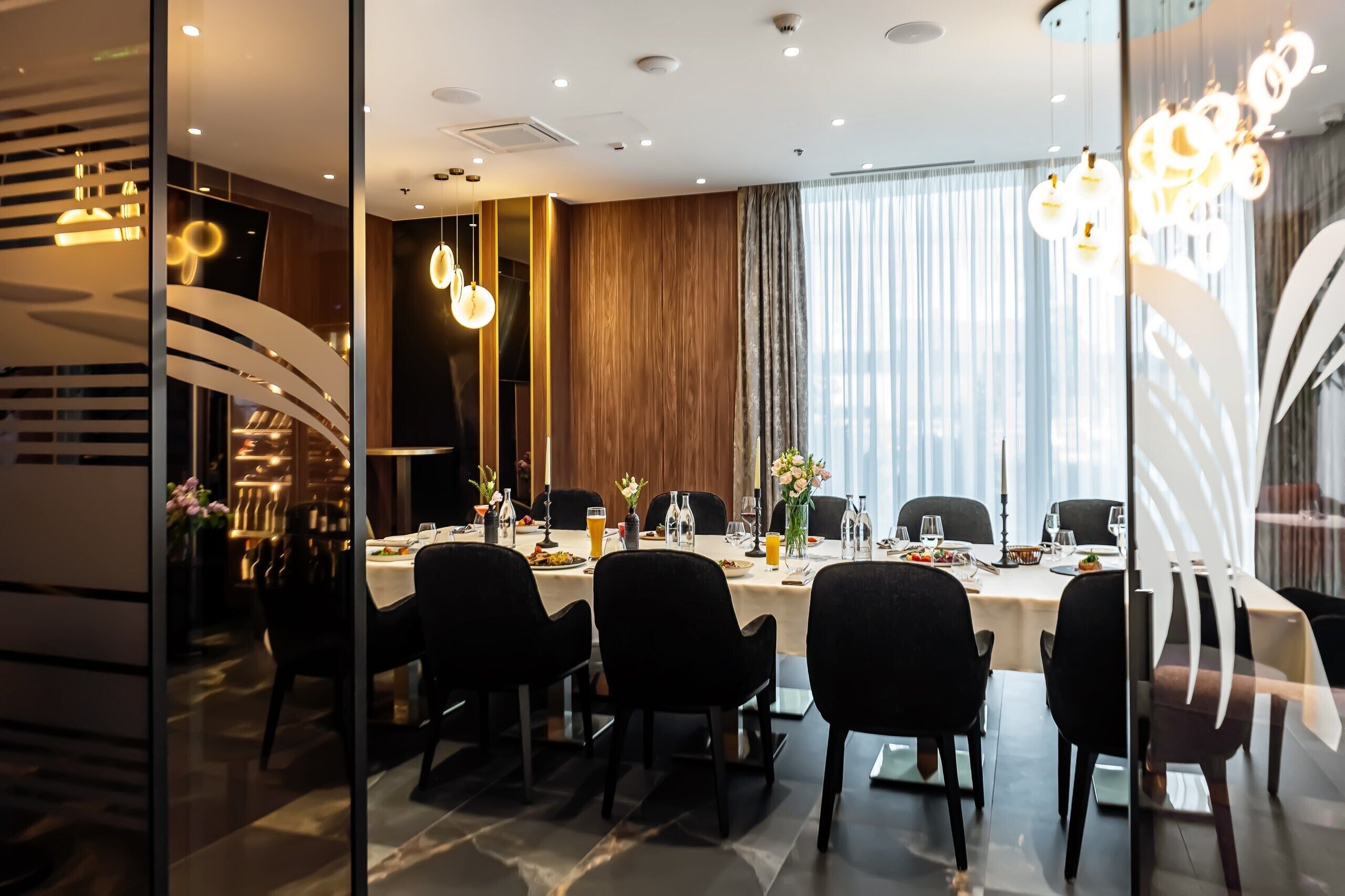 A modern dining area with a long table, black chairs, and a glass partition.