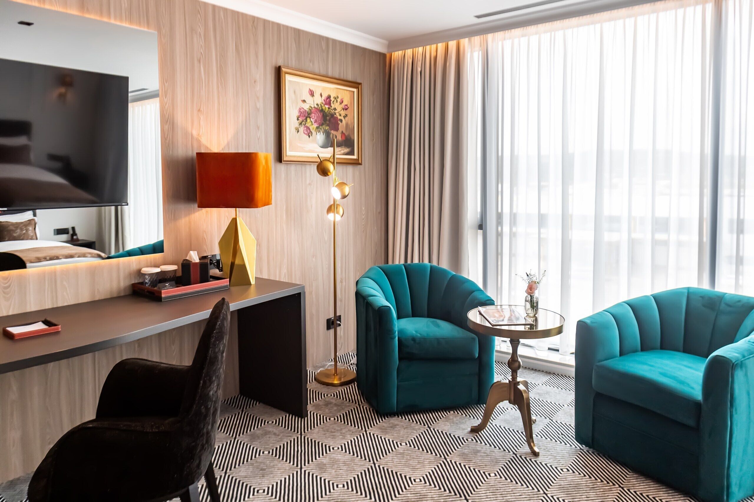 A hotel room with a bed, a desk with a TV, two teal armchairs, a floor lamp, and a large window with curtains.
