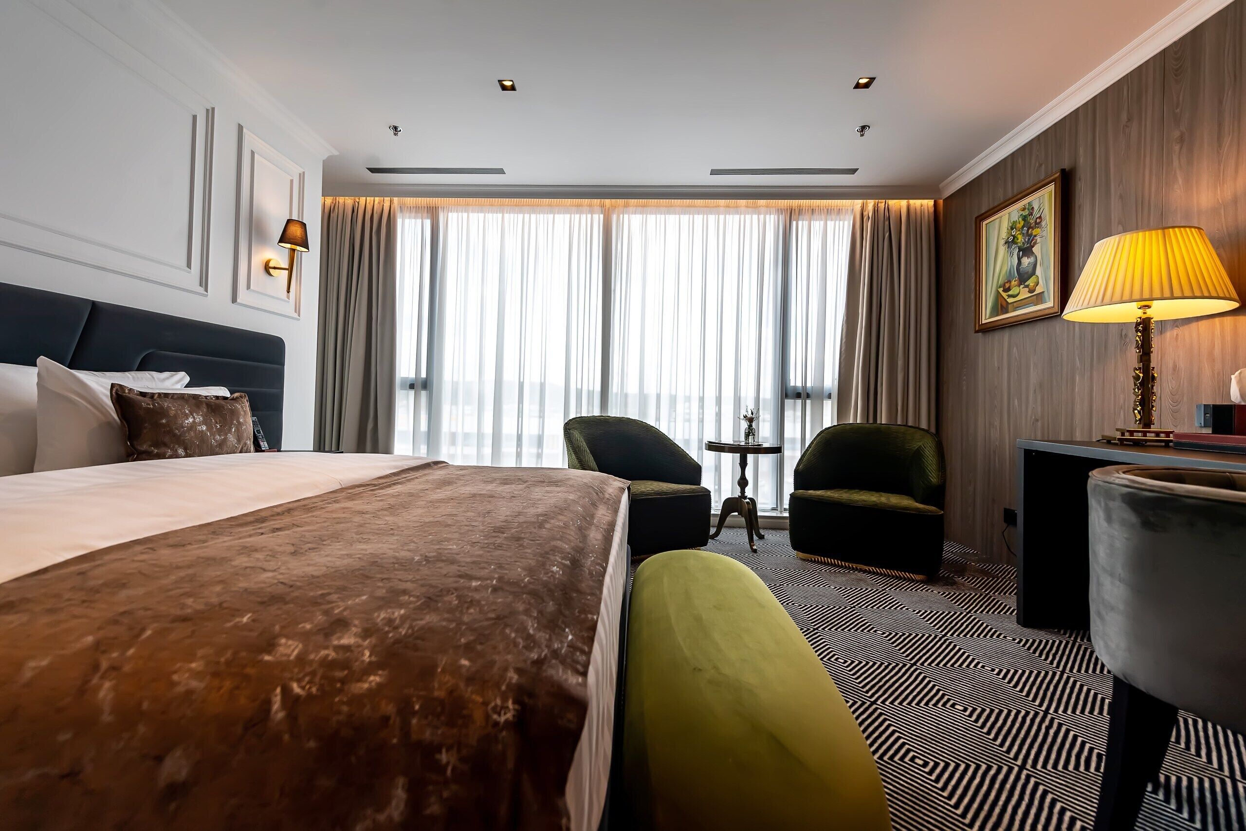 Habitación de hotel con una cama grande, un escritorio, una silla, una lámpara y vistas a la ciudad a través de amplias ventanas.