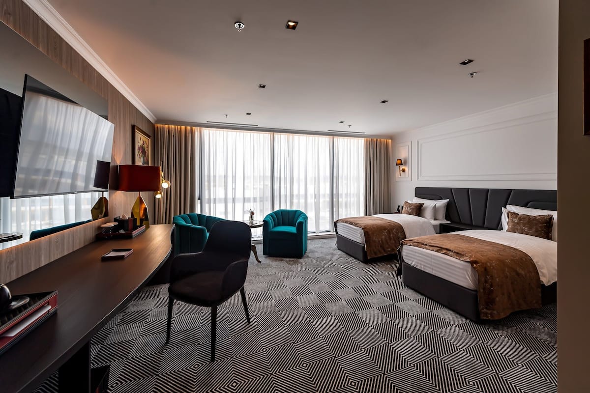 Habitación de hotel con escritorio, dos camas, televisor y un ventanal grande con cortinas.