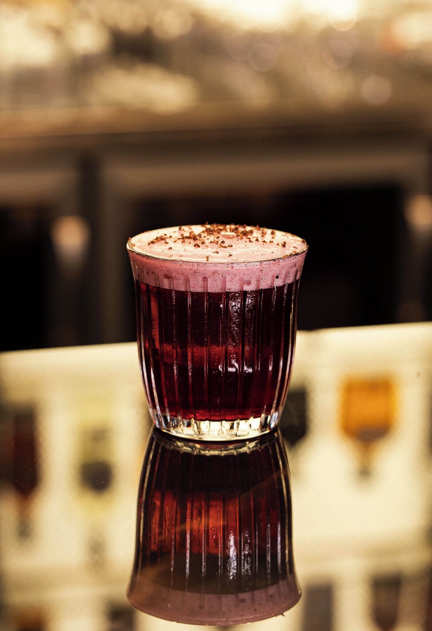 A glass of dark-colored beverage with a frothy top, placed on a reflective surface.