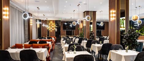Zona pranzo di un hotel con diversi tavoli, sedie e un grande televisore a schermo piatto.