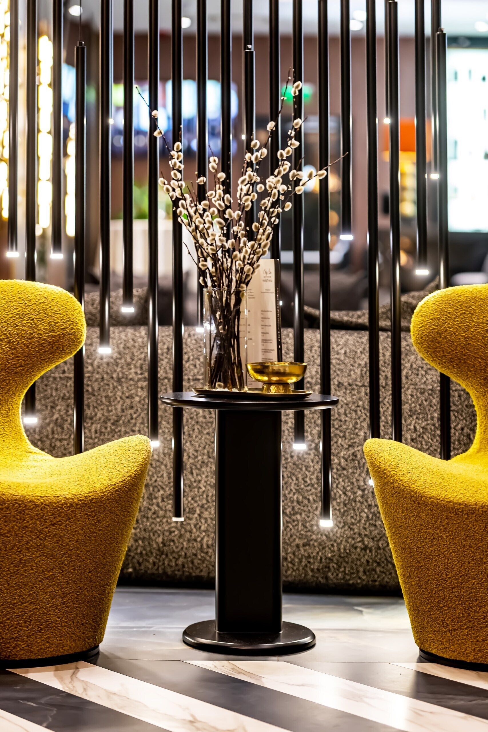 Un intérieur moderne avec deux chaises jaunes, une table noire ornée d’un vase rempli de branches et un mur décoratif aux lignes verticales.