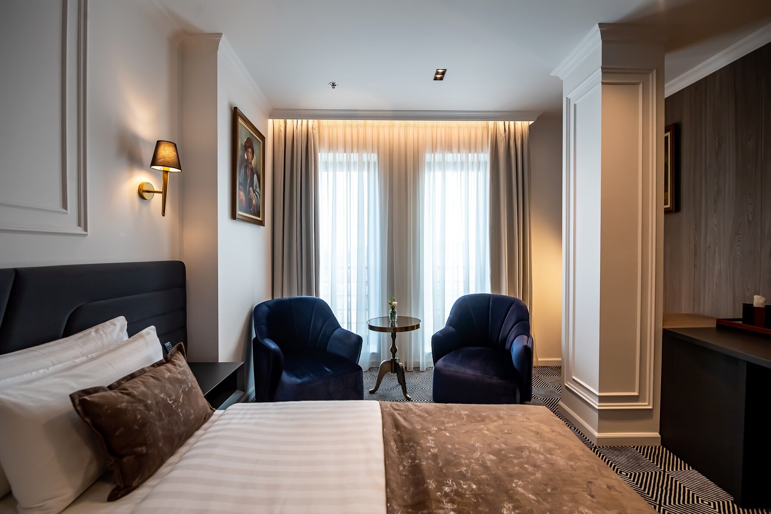 A hotel room with a bed, two armchairs, a small table, and a view of the outdoors through large windows.