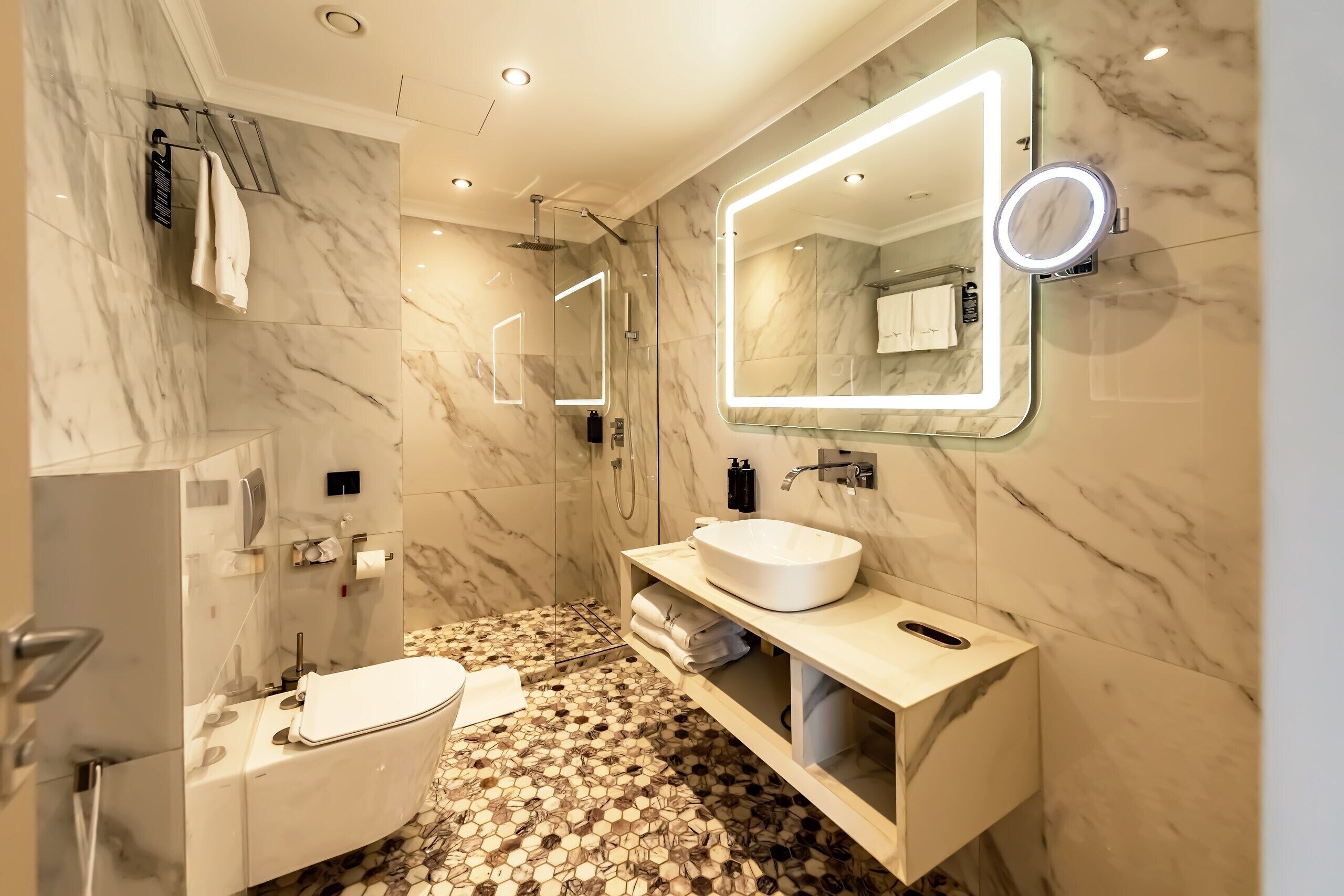 Un baño moderno con suelo de mármol, una bañera independiente, un tocador con lavabo y un espejo grande con iluminación integrada.