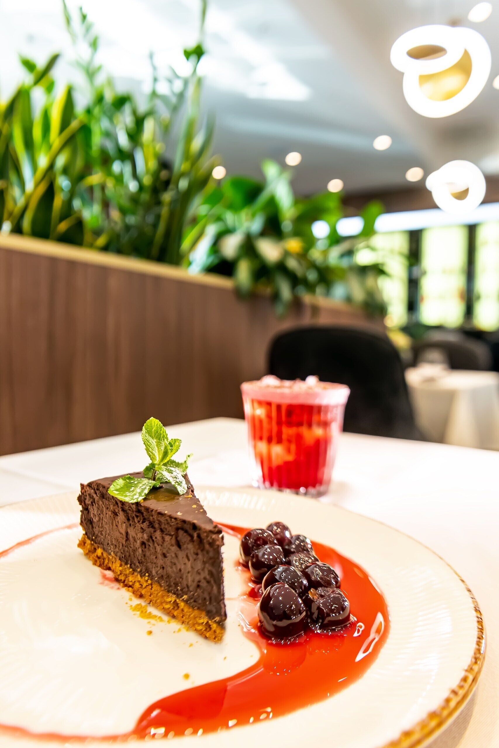 En dessert garnerad med mynta, serverad på en tallrik tillsammans med ett glas röd dryck, placerad på ett bord i en restaurang med krukväxter i bakgrunden.