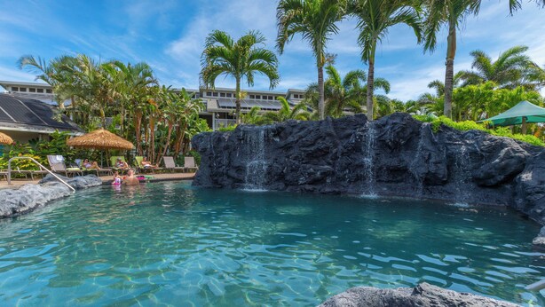 A pool with a waterfall feature, surrounded by palm trees and lounge chairs. - Princeville condo with ocean views, pool and hot tub access summer deals (PRINCEVILLE)