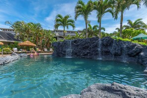 A pool with a waterfall feature, surrounded by palm trees and lounge chairs. - Princeville condo with ocean views, pool and hot tub access summer deals (PRINCEVILLE)