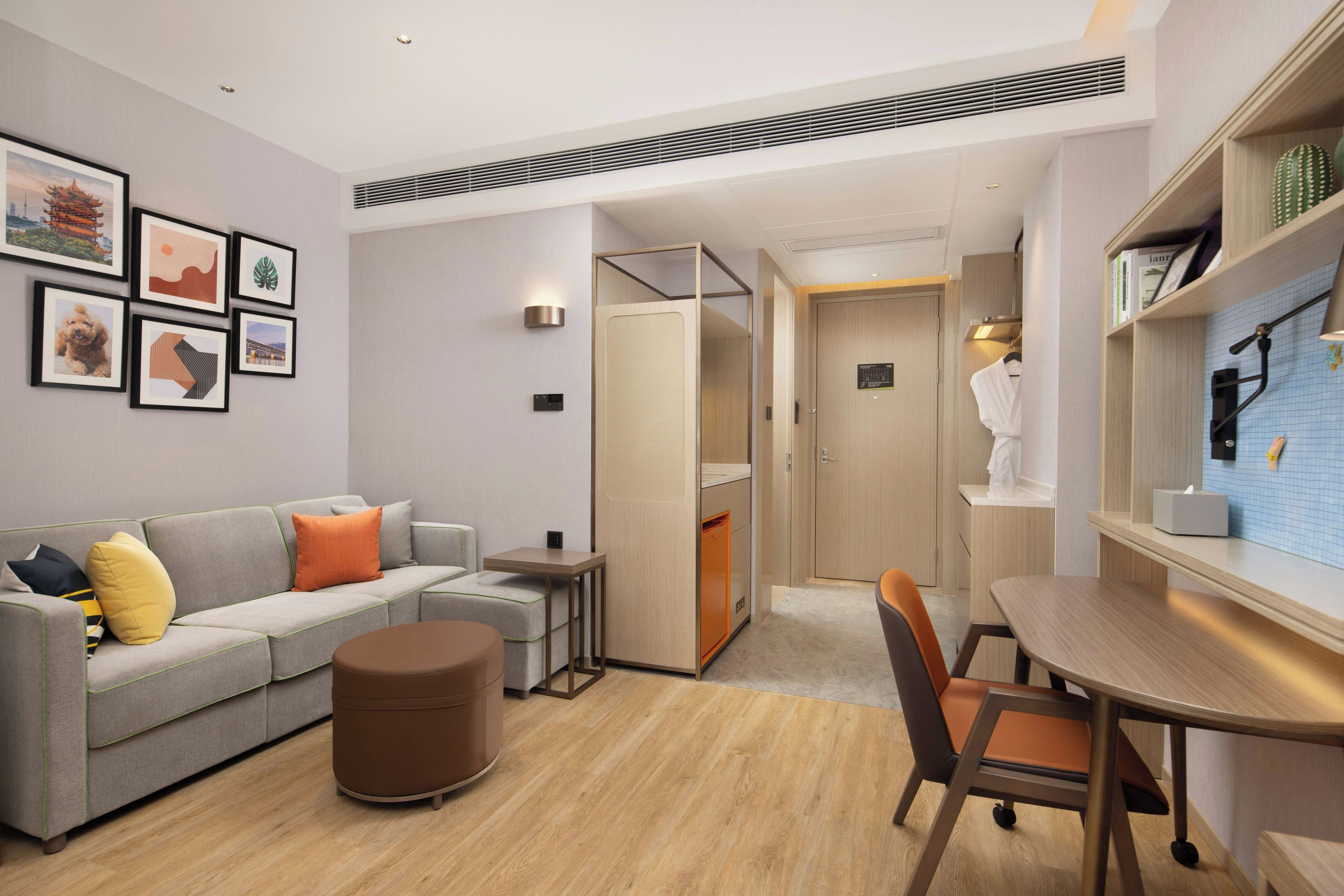 Ein modernes Hotelzimmer mit grauem Sofa, einem kleinen runden Hocker, einem Holztisch und einem Badezimmer mit Handtuchhalter.