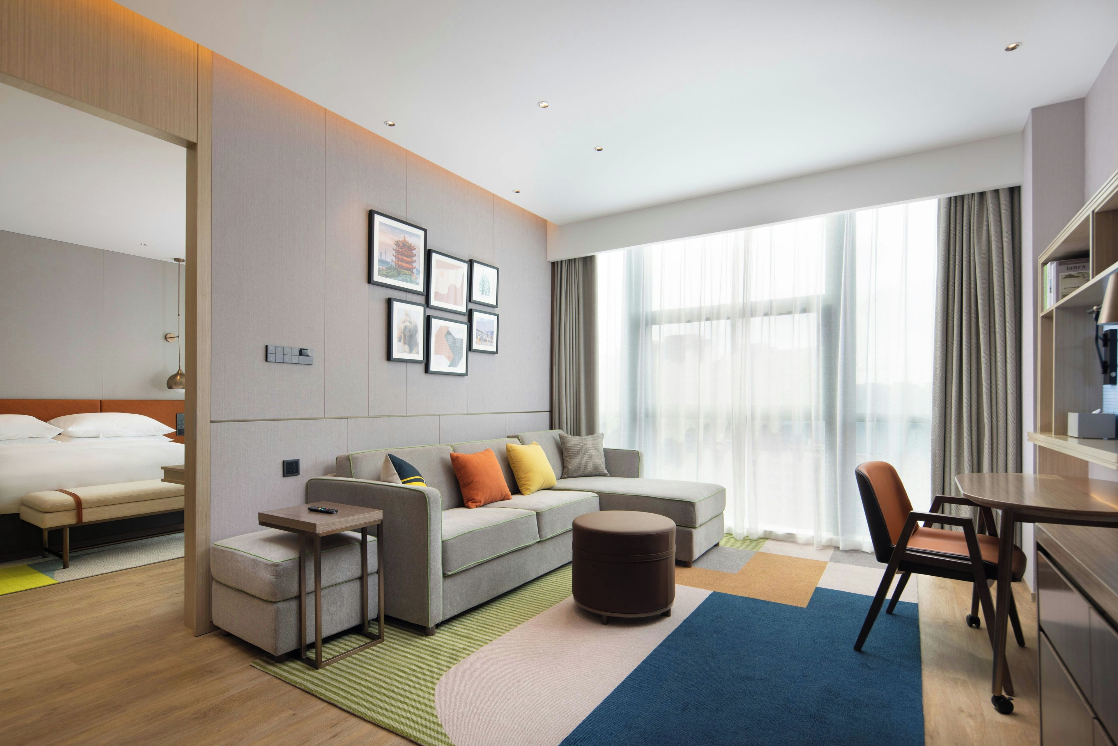 A hotel room with a bed, a sofa, a coffee table, a chair, and a desk. There are framed pictures on the wall, a window with curtains, and a large window with a view.