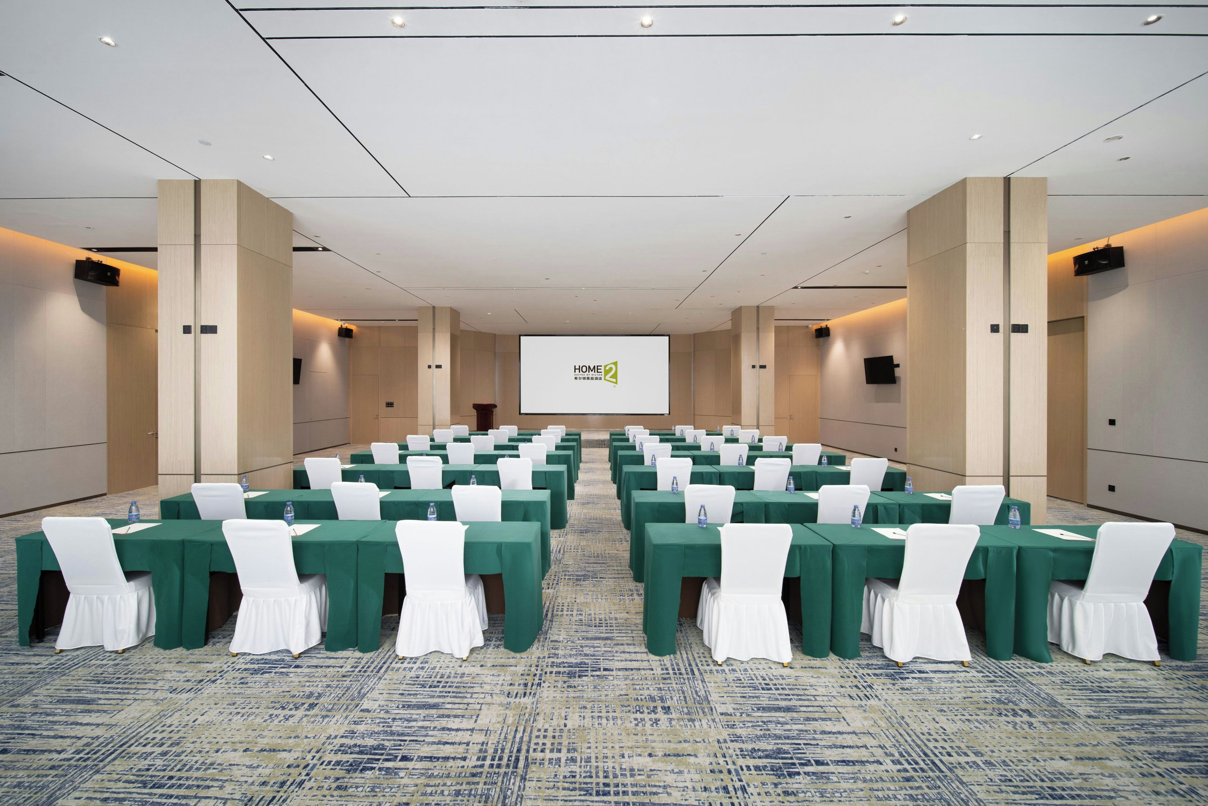 Un salón de conferencias con filas de mesas, sillas y una pantalla grande al frente.