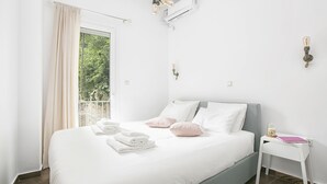 Een keurig opgemaakt bed met witte en roze kussens, een klein nachtkastje met een notitieboekje, en een airconditioner aan de muur.