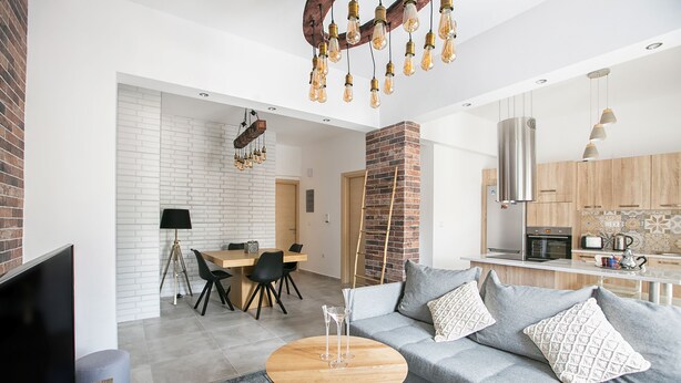 Een moderne woonkamer met een grijze bank, een houten salontafel en een televisie. Er is een zithoek met een tafel en stoelen, en een keuken met houten kasten en een fornuis.
