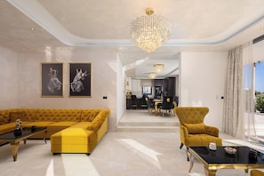 A spacious living room with a yellow sectional sofa, a black coffee table, and a yellow armchair. There are two framed artworks on the wall, a chandelier, and a view of the kitchen in the background.