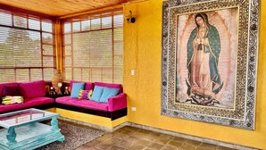 A room with a wooden ceiling, a yellow wall, a large framed religious painting, a pink sofa, and a glass coffee table.
