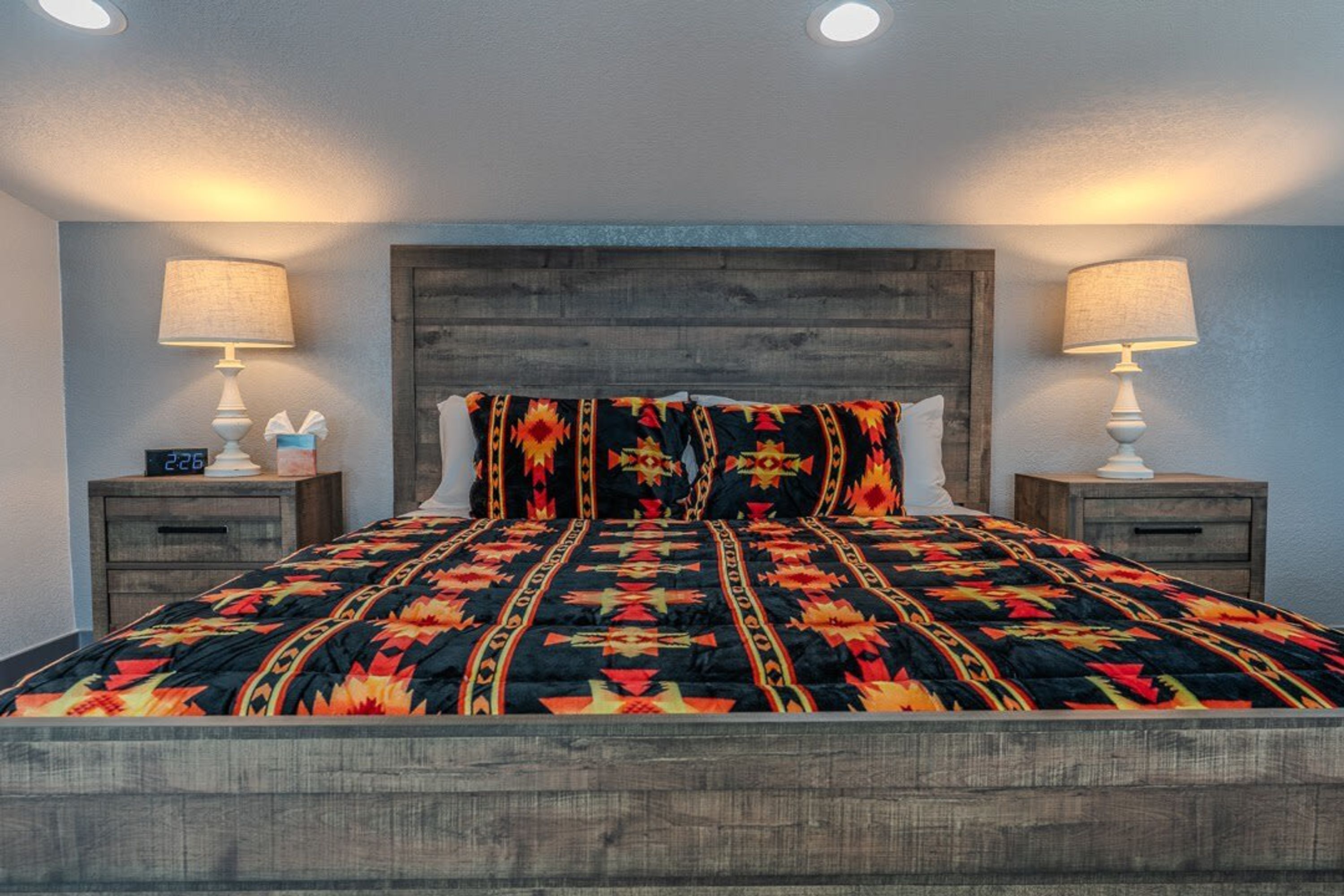 Una cama con un edredón estampado, dos mesitas de noche con lámparas y un cabecero de madera.