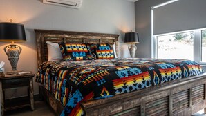 Un dormitorio con estructura de cama de madera, colcha colorida, dos lámparas de noche y una ventana con vistas al exterior.