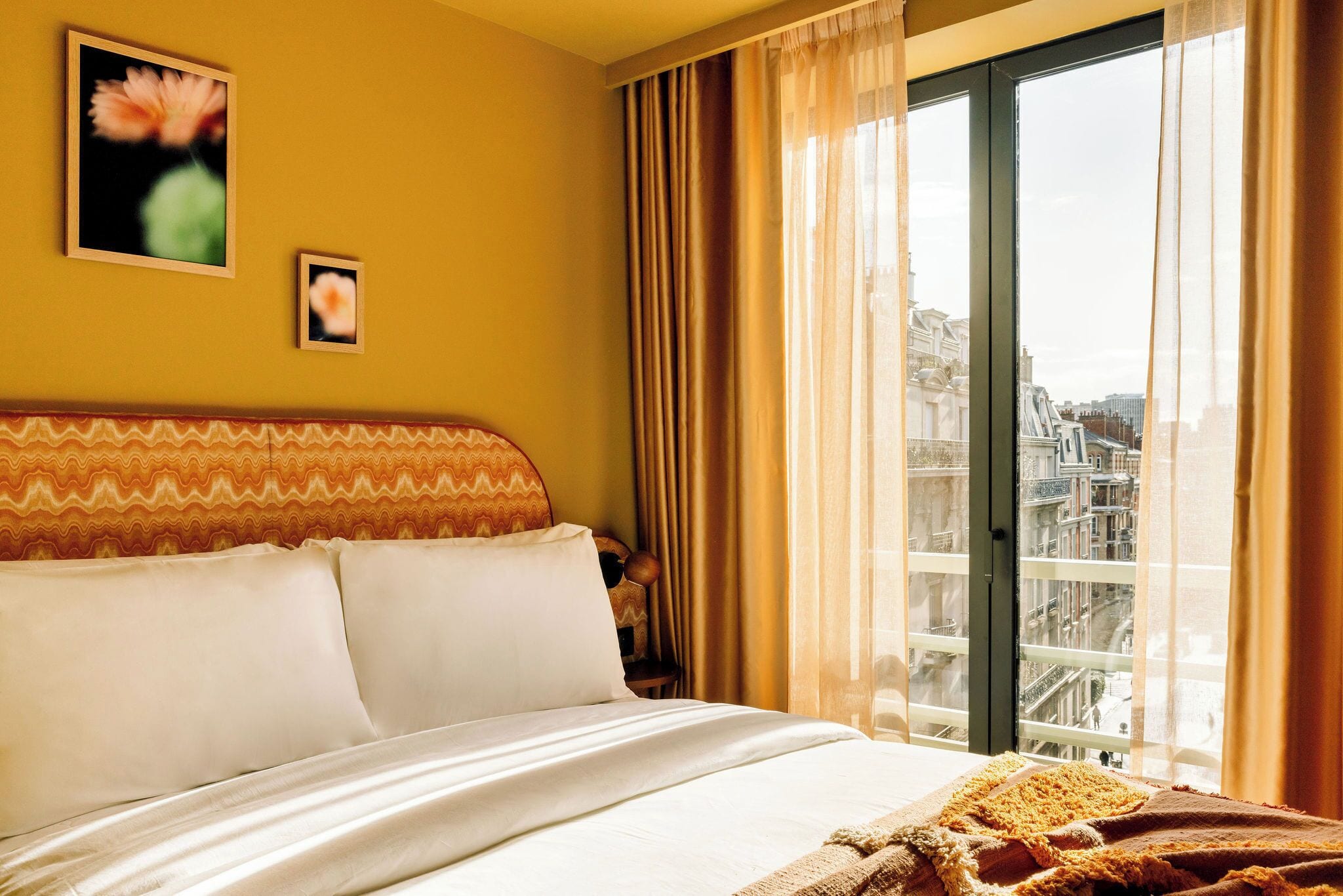Ein Hotelzimmer mit Bett, Blick auf die Stadt durch ein großes Fenster und einer Wand mit zwei gerahmten Bildern.