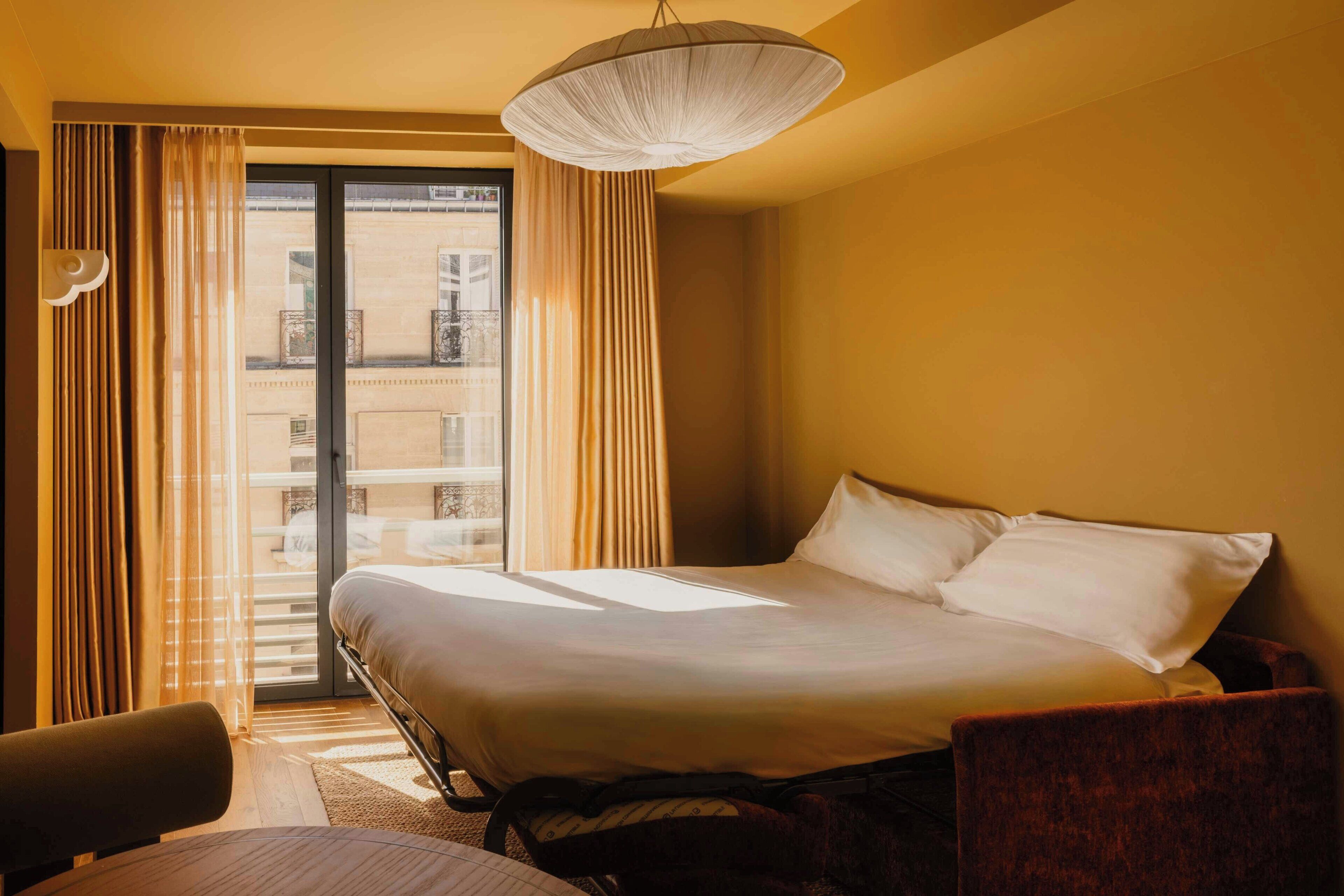 A hotel room with a bed, a chair, a desk, a large window with curtains, and a view of the building outside.