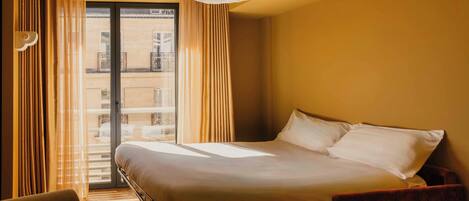 A hotel room with a bed, a chair, a desk, a large window with curtains, and a view of the building outside.