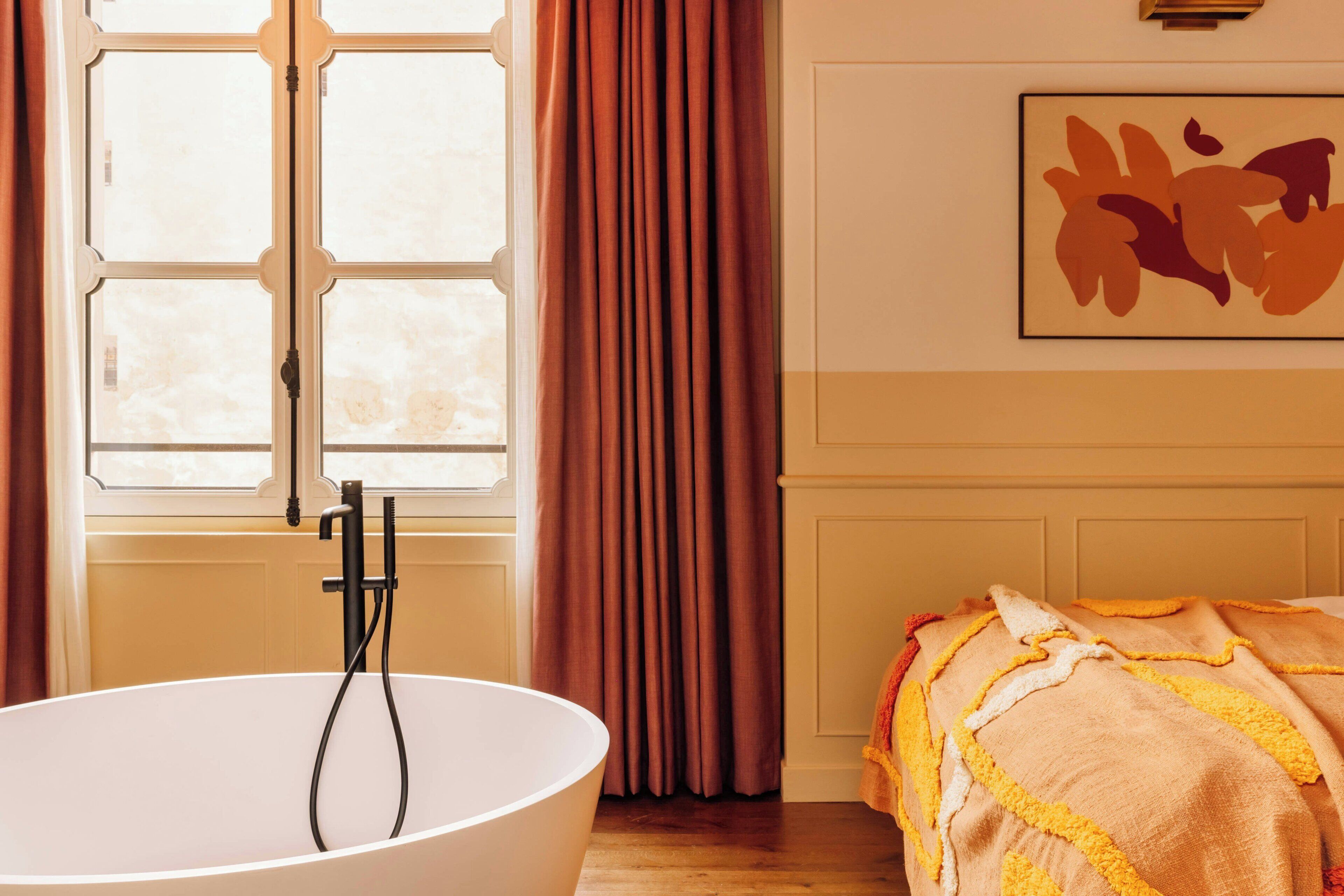 A room with a large bathtub, a window with curtains, and a bed with a blanket.