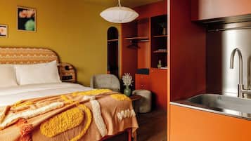 A bedroom with a bed, a bedside table, and a nightstand. There is a small seating area with a chair and a stool. The room has a red wall and a wooden floor.
