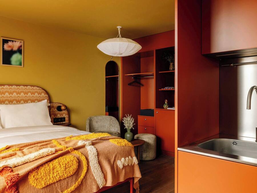 A bedroom with a bed, a bedside table, and a nightstand. There is a small seating area with a chair and a stool. The room has a red wall and a wooden floor.