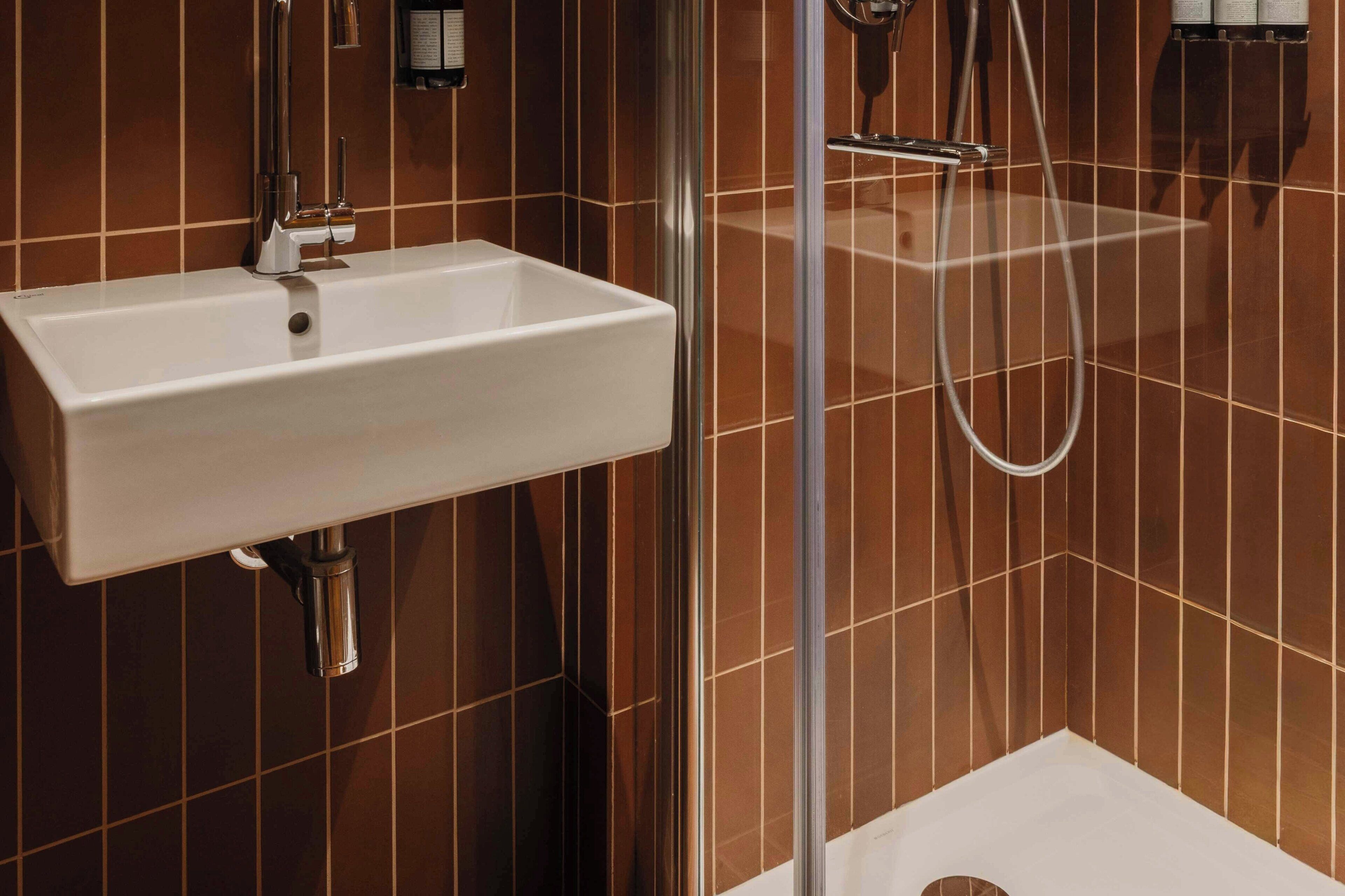 Un baño con lavamanos blanco, ducha y paredes de azulejos marrones.