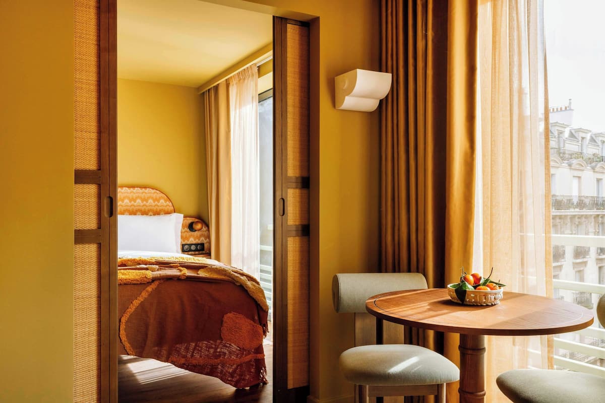 Hotellihuone, jossa on sänky, pyöreä puinen pöytä, jonka päällä on kulho täynnä hedelmiä, ja tuoli.