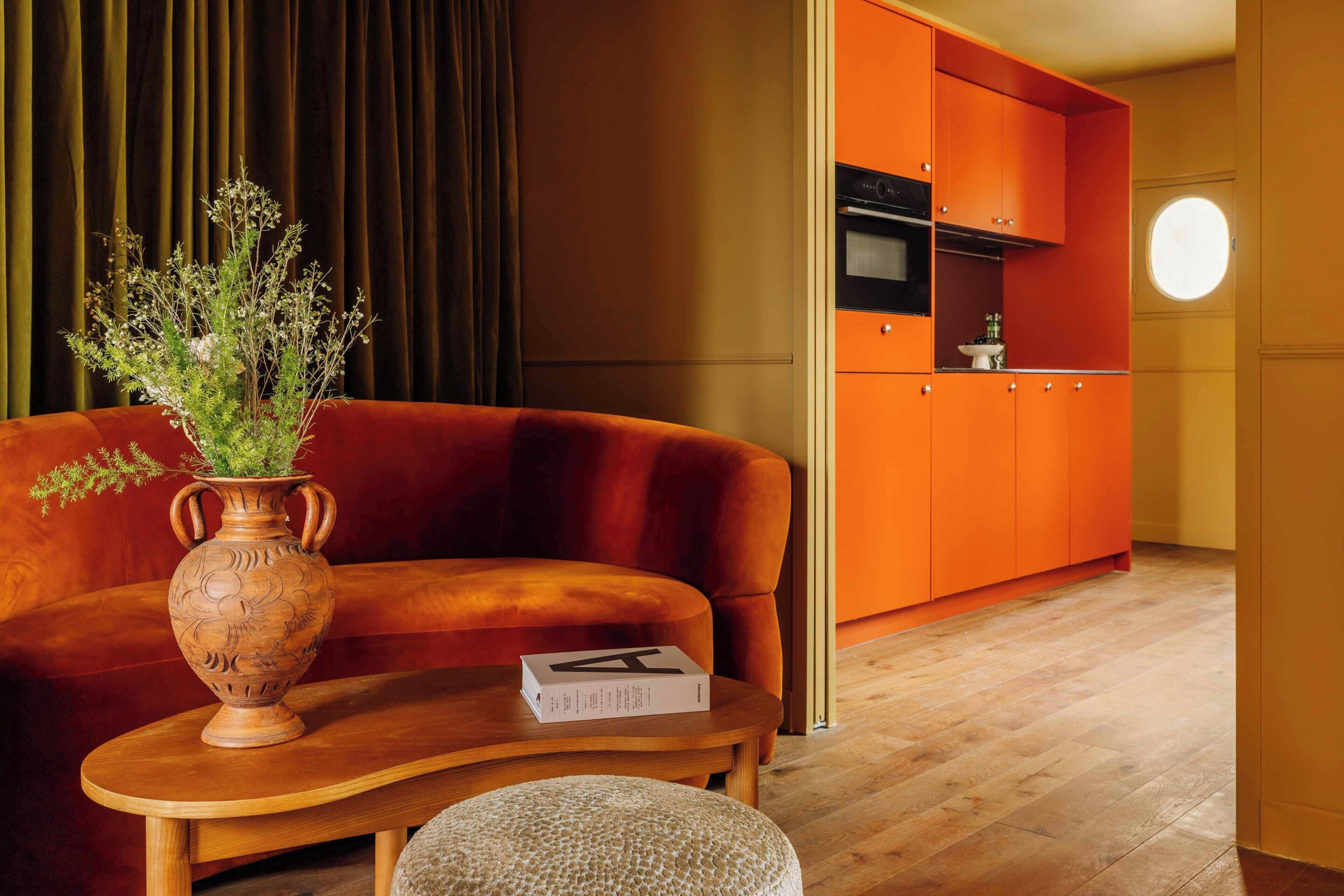 A modern living room with a sofa, a wooden coffee table, a vase with flowers, and a kitchen area with orange cabinets and a round window.