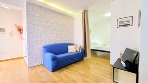 Une pièce avec un canapé bleu, un lit recouvert de linge de lit blanc, un plancher en bois, un mur à l’aspect pierre et un tableau encadré accroché au mur.