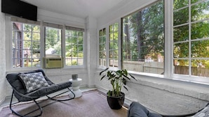A sunlit room with large windows, a black rocking chair, a gray sofa, and a potted plant.