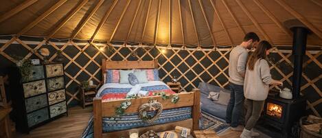 A cozy interior of a yurt with a bed, a small table with items, a fireplace, and a lattice wall.