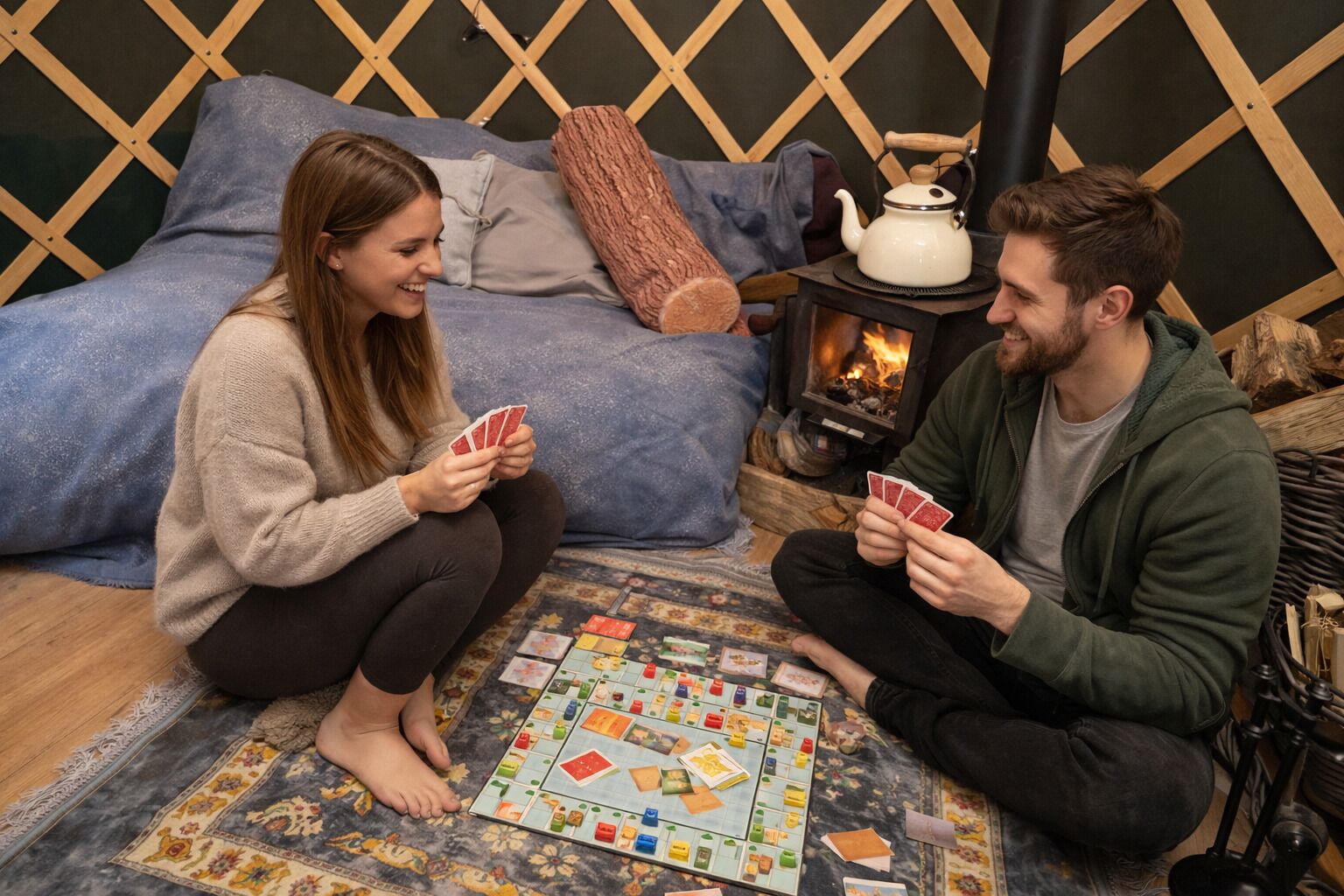Deux personnes jouent à un jeu de société sur un tapis à motifs dans une pièce où se trouvent un lit, des coussins et un poêle à bois.