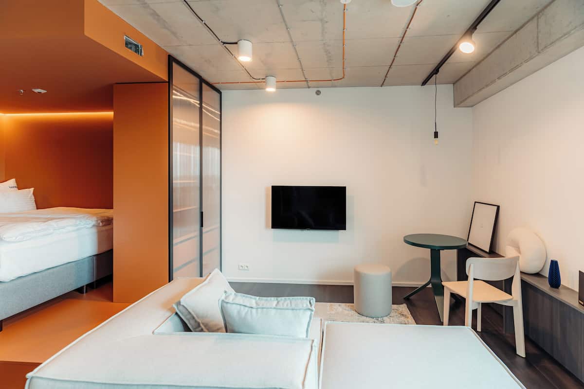 A modern hotel room with a bed, a sofa, a small table, and a TV mounted on the wall.